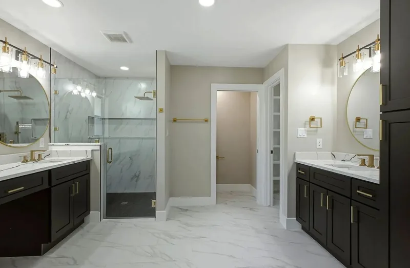 Master bathroom with twin dark vanities, marble effect tile, round gold framed mirrors and a glass shower
