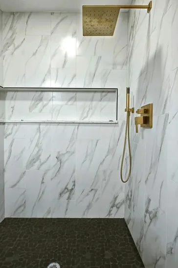 Walk in shower in marble effect tile with a gold rain head, recessed shelf and a dark pebble floor