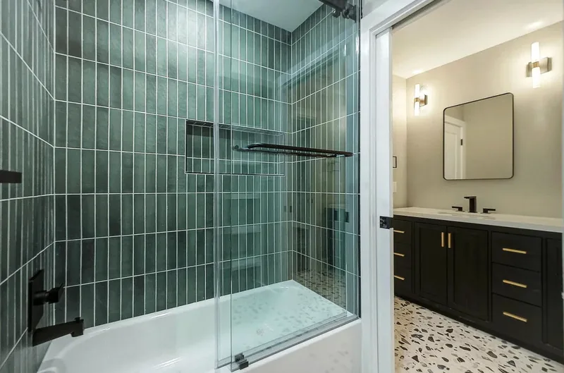 Tub and shower in dark green vertical tile, with a terrazzo floor and dark vanity beyond